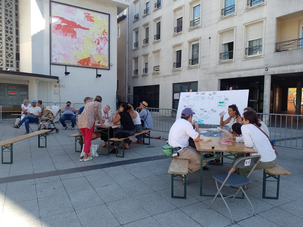Place du théâtre, atelier 2030 Glorieuses : 2 tables où les gens discutent et 1 panneau pour la facilitation graphique (dessins)