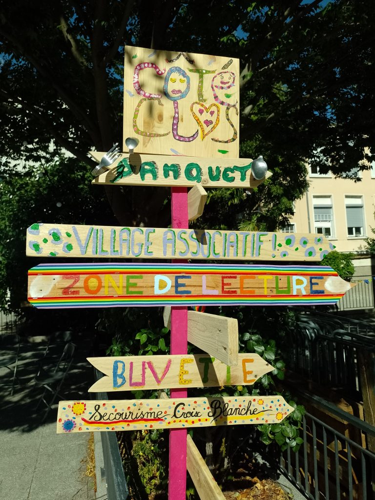 Place du théâtre : panneau indicatifs colorés, en bois peint et décoré par les bénévoles d'arc en ciel, de visages, de fourchettes et cuiller tordues 